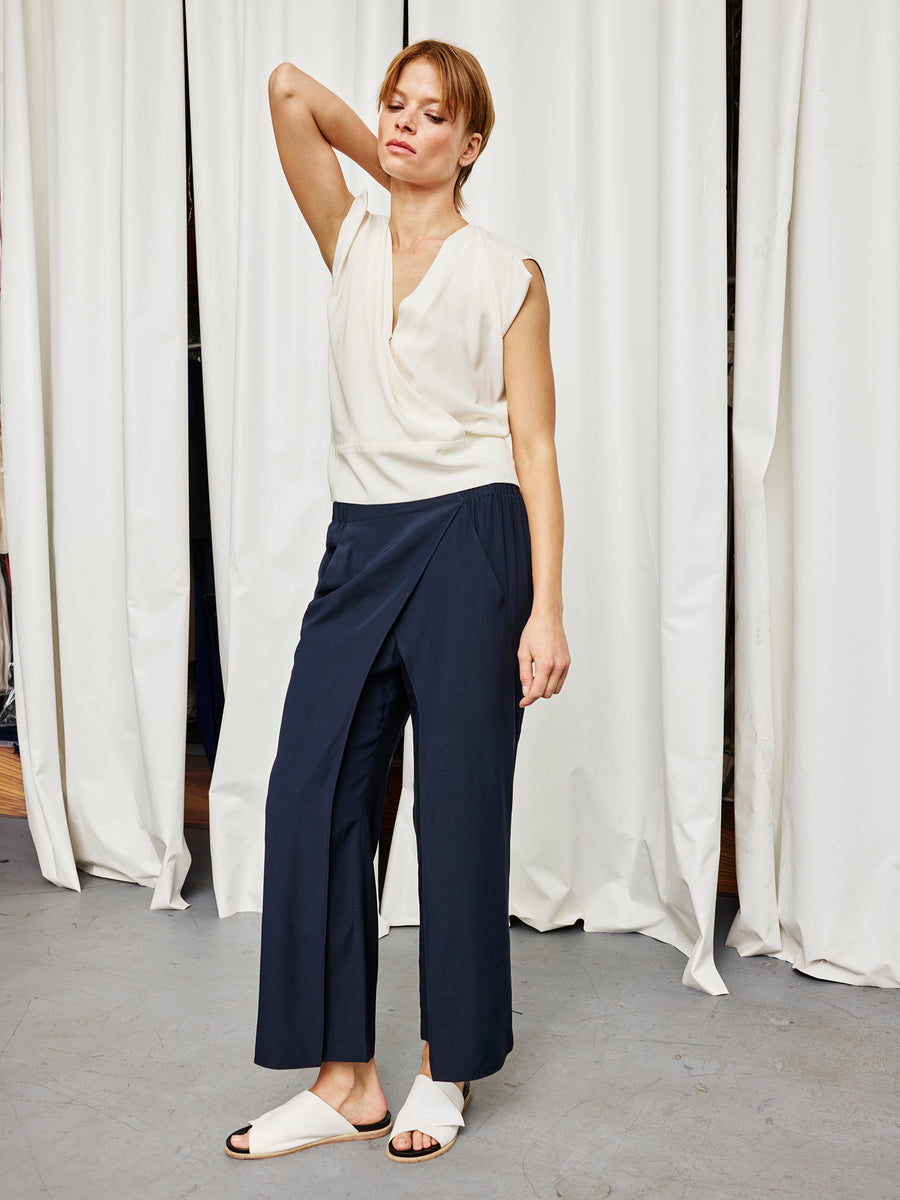A woman in front of white curtains wears the Ido Pant by Zero + Maria Cornejo, a sleeveless white blouse, and white slide sandals. She poses with one arm raised and looks relaxed. Made in New York.