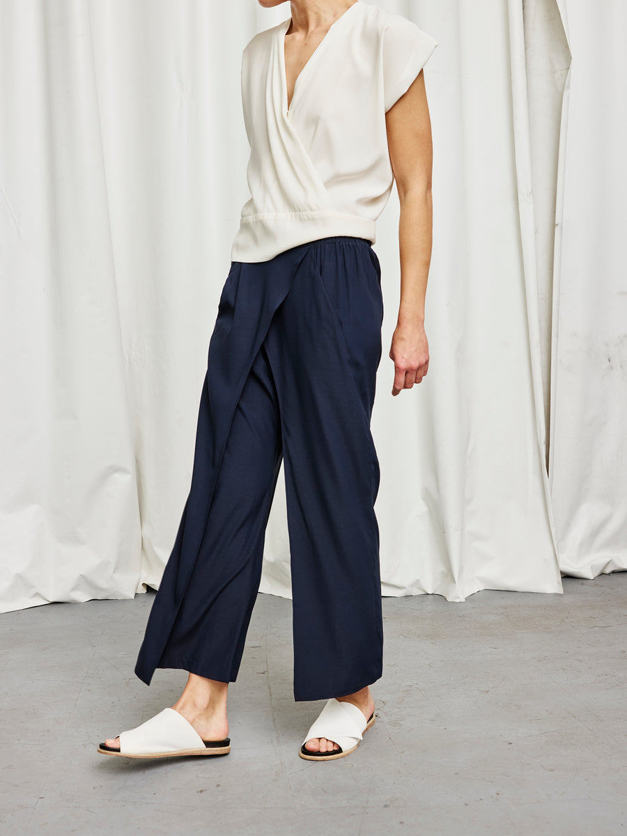 A person wears a white sleeveless blouse, Zero + Maria Cornejo Ido Pant in navy wide-leg stretch silk, and white slide sandals, standing on a gray floor with white curtains behind. Their face is out of frame.