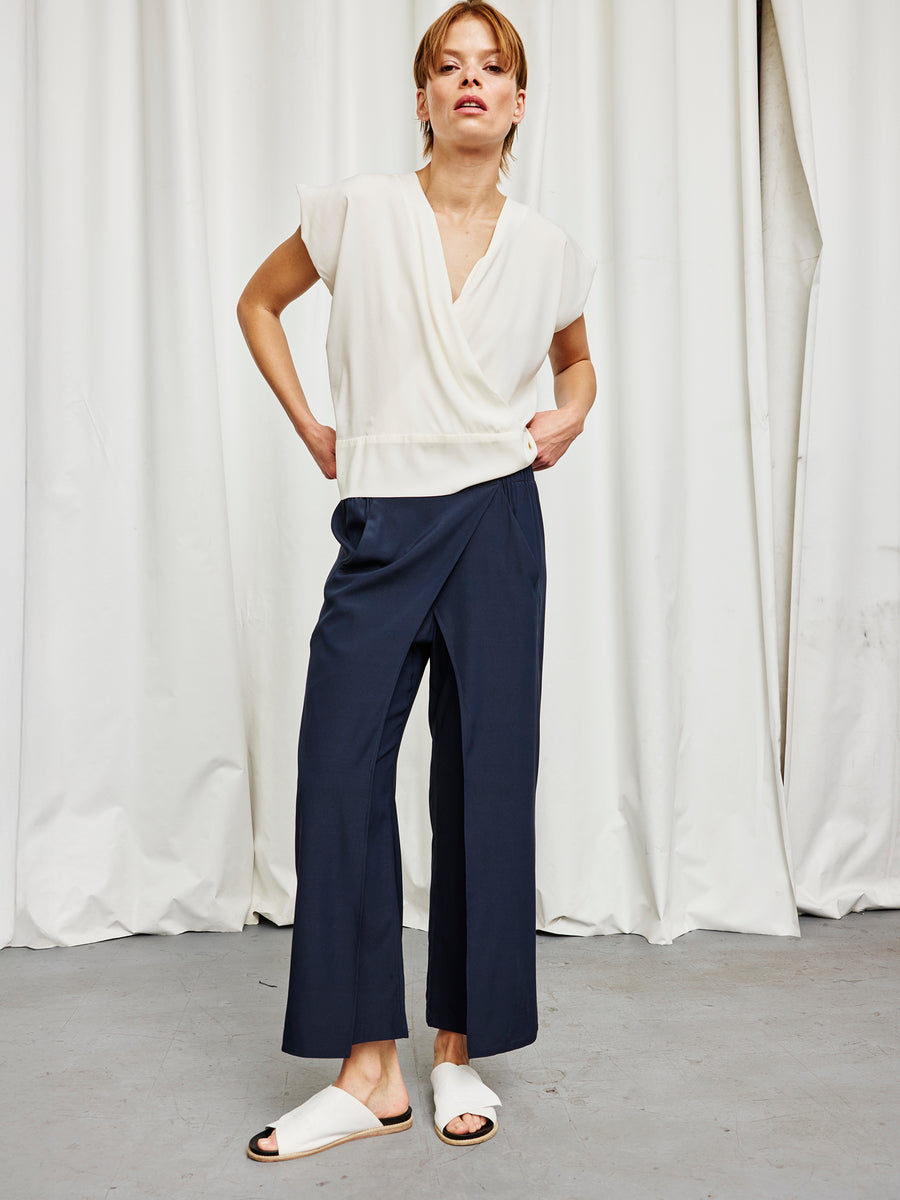 A person stands before white curtains wearing a white wrap top, the Zero + Maria Cornejo Ido Pant in navy silk charmeuse, and white slide sandals—an elegant New York-made look with a neutral expression and hands on hips.