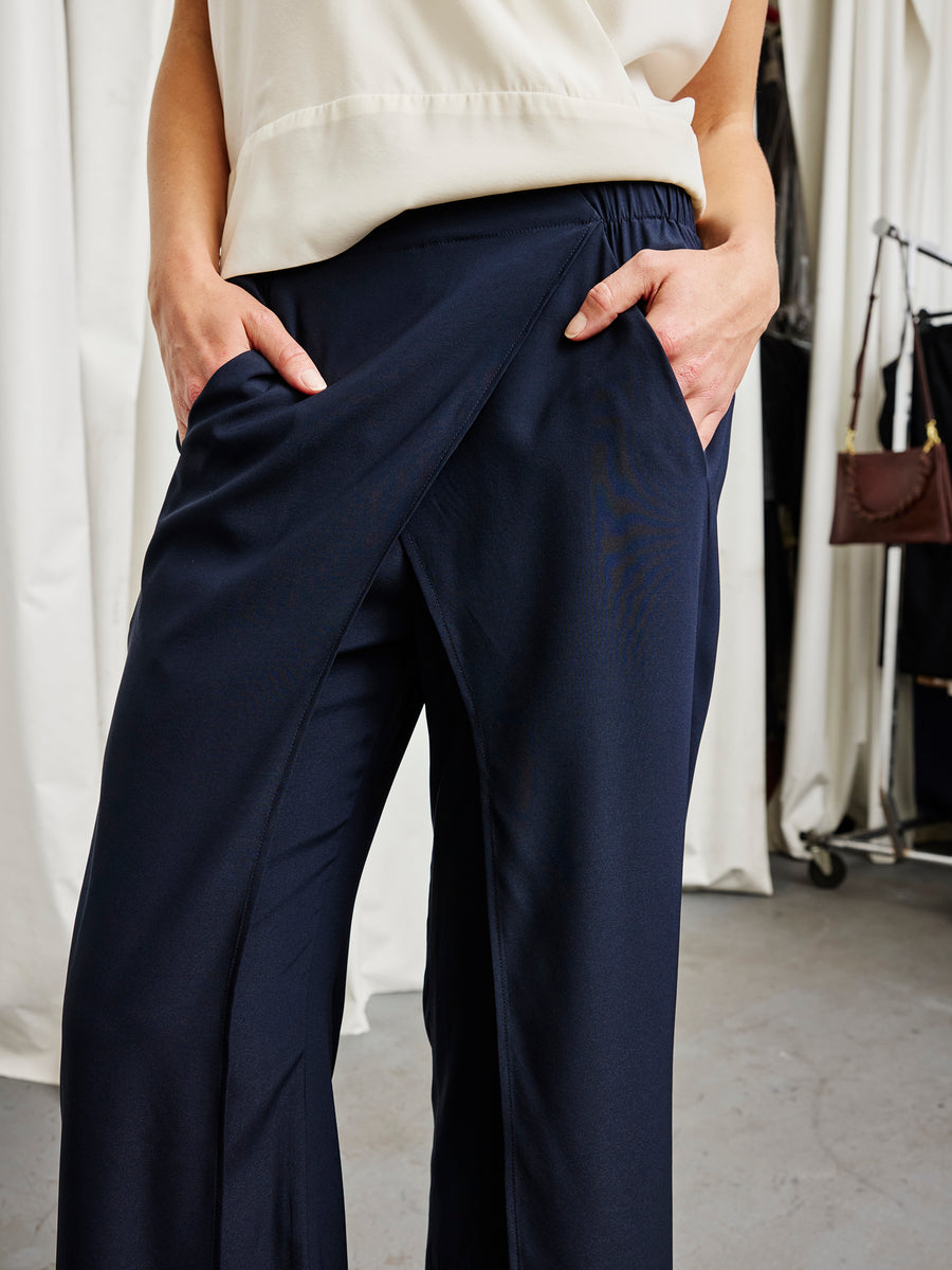 A person wearing Zero + Maria Cornejo's Ido Pant in navy stretch silk with a cream top stands with hands in pockets, in front of white curtains and a clothing rack.