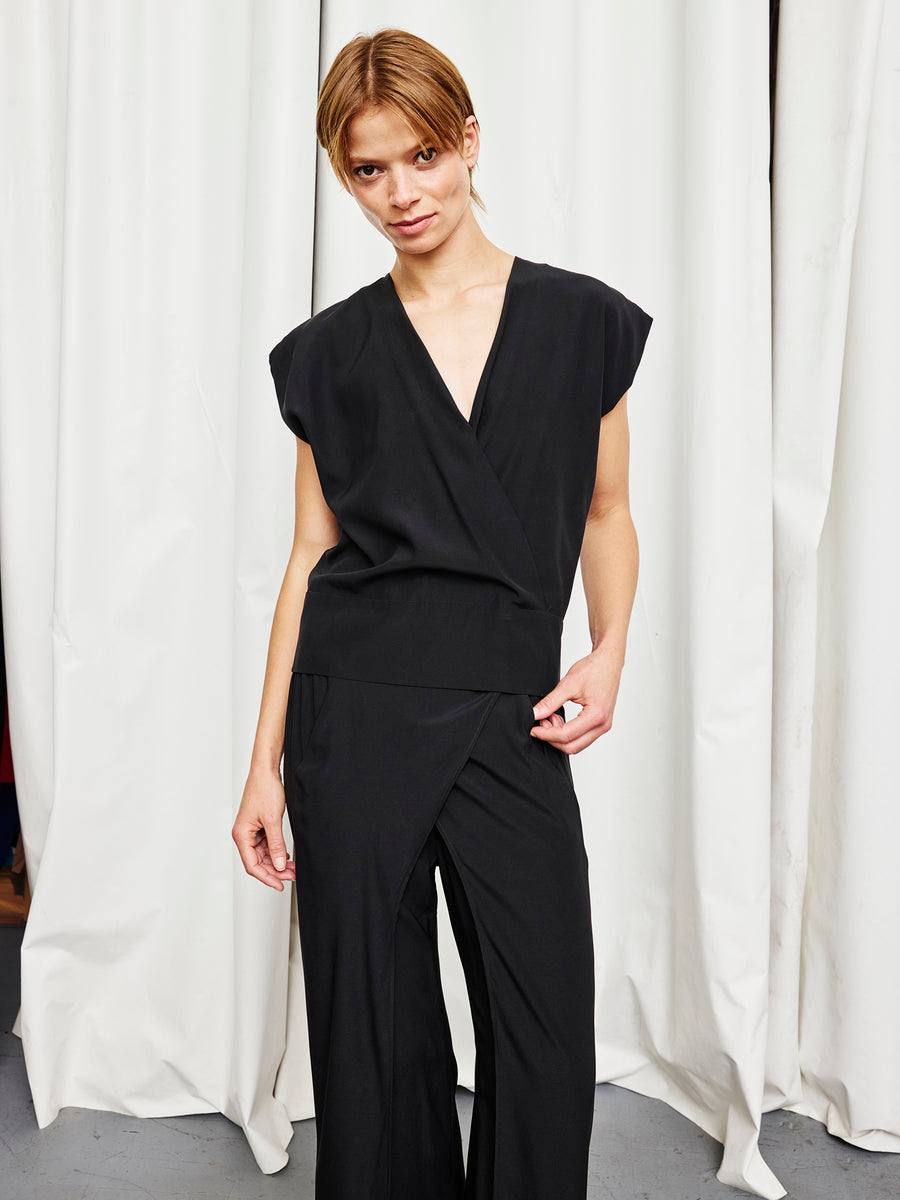 A person stands before white curtains, wearing the Zero + Maria Cornejo Wrap Mido Top—a sleeveless black piece with a deep V-neck—paired with wide-leg pants, looking at the camera with a neutral expression; made in New York.