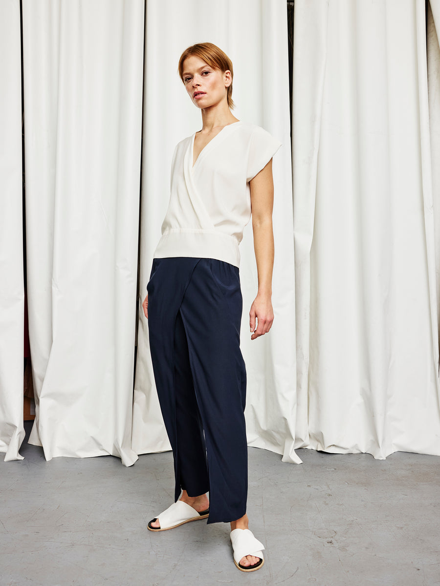 A person stands before white curtains in the Zero + Maria Cornejo Wrap Mido Top, loose navy blue pants, and white slide sandals—made in New York—looking confidently at the camera.