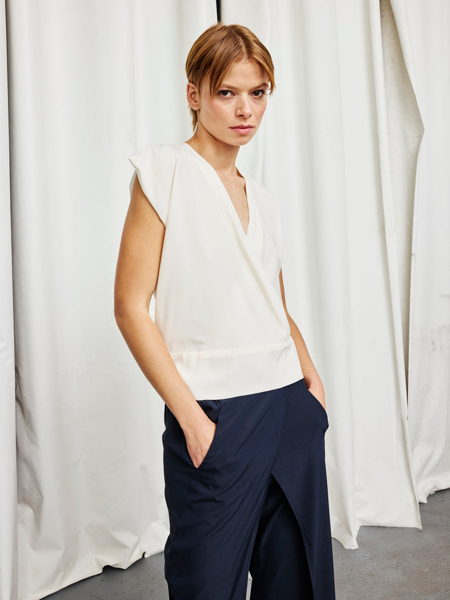 A young woman stands before white curtains, wearing the Zero + Maria Cornejo Wrap Mido Top and navy blue pants, with her hands in her pockets and a neutral expression as she looks at the camera.