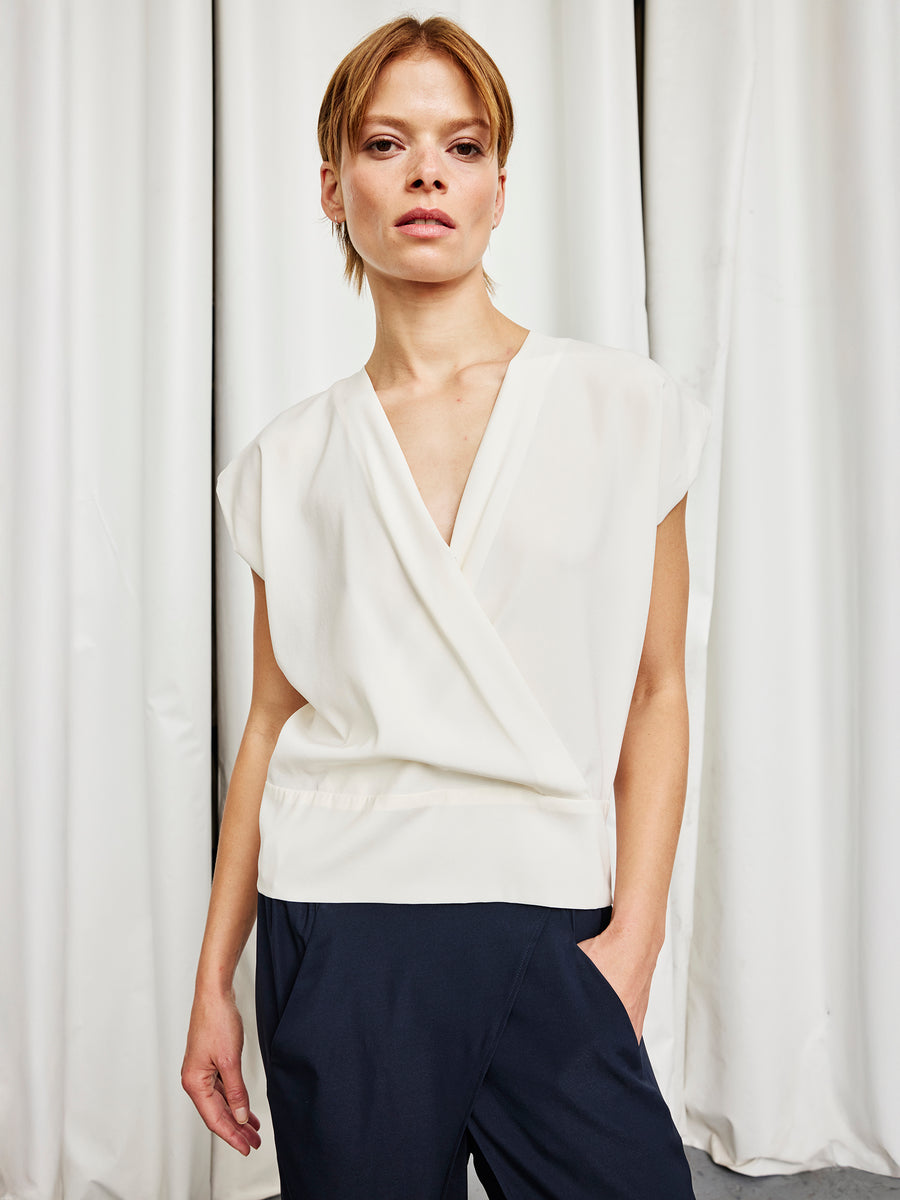 A person with short, light brown hair wears the Zero + Maria Cornejo Wrap Mido Top in white silk charmeuse and dark pants, standing before white curtains with one hand in their pocket, looking confidently at the camera. Made in New York.