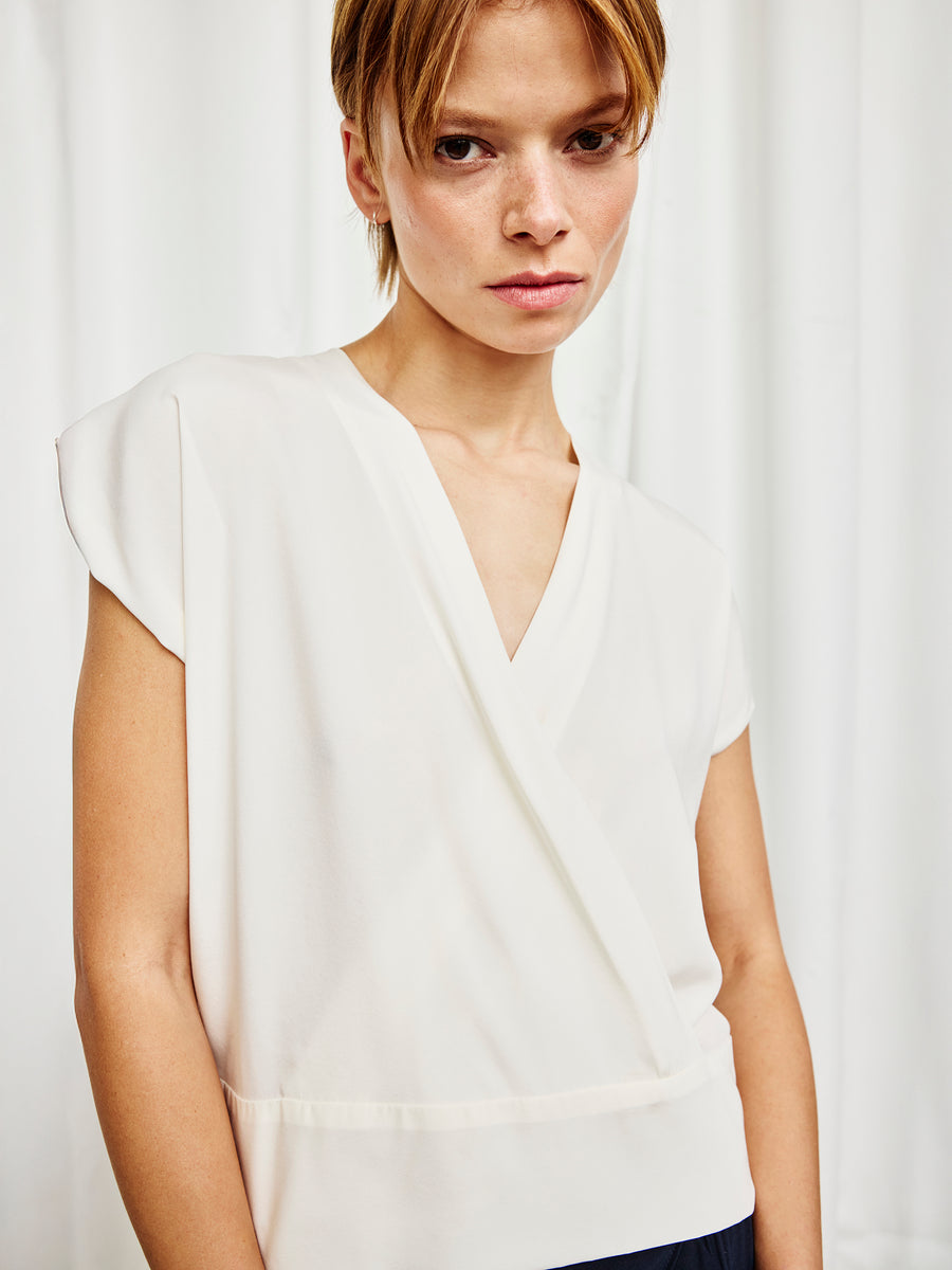 A person with short light brown hair wears the Zero + Maria Cornejo Wrap Mido Top, featuring a white silk charmeuse V-neck wrap design, standing confidently before white curtains and looking at the camera.
