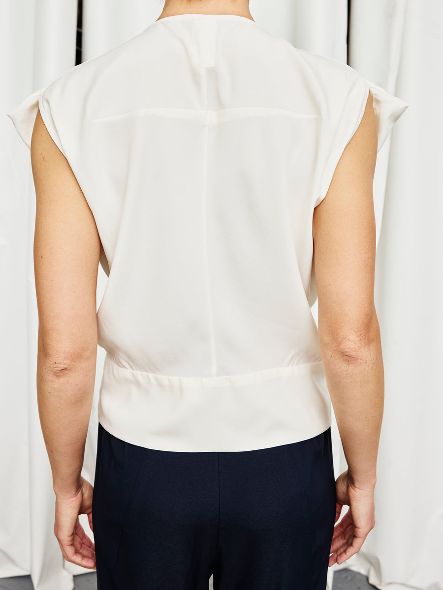 A person is seen from behind, wearing the Zero + Maria Cornejo Wrap Mido Top and dark pants, standing in front of white curtains.