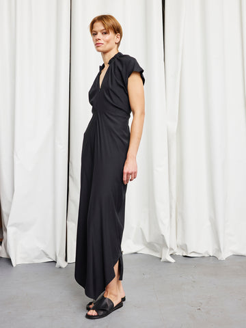 A woman with short hair stands before white curtains, wearing the Zero + Maria Cornejo Long Ruched Stella Dress and black sandals. She looks slightly to the side.