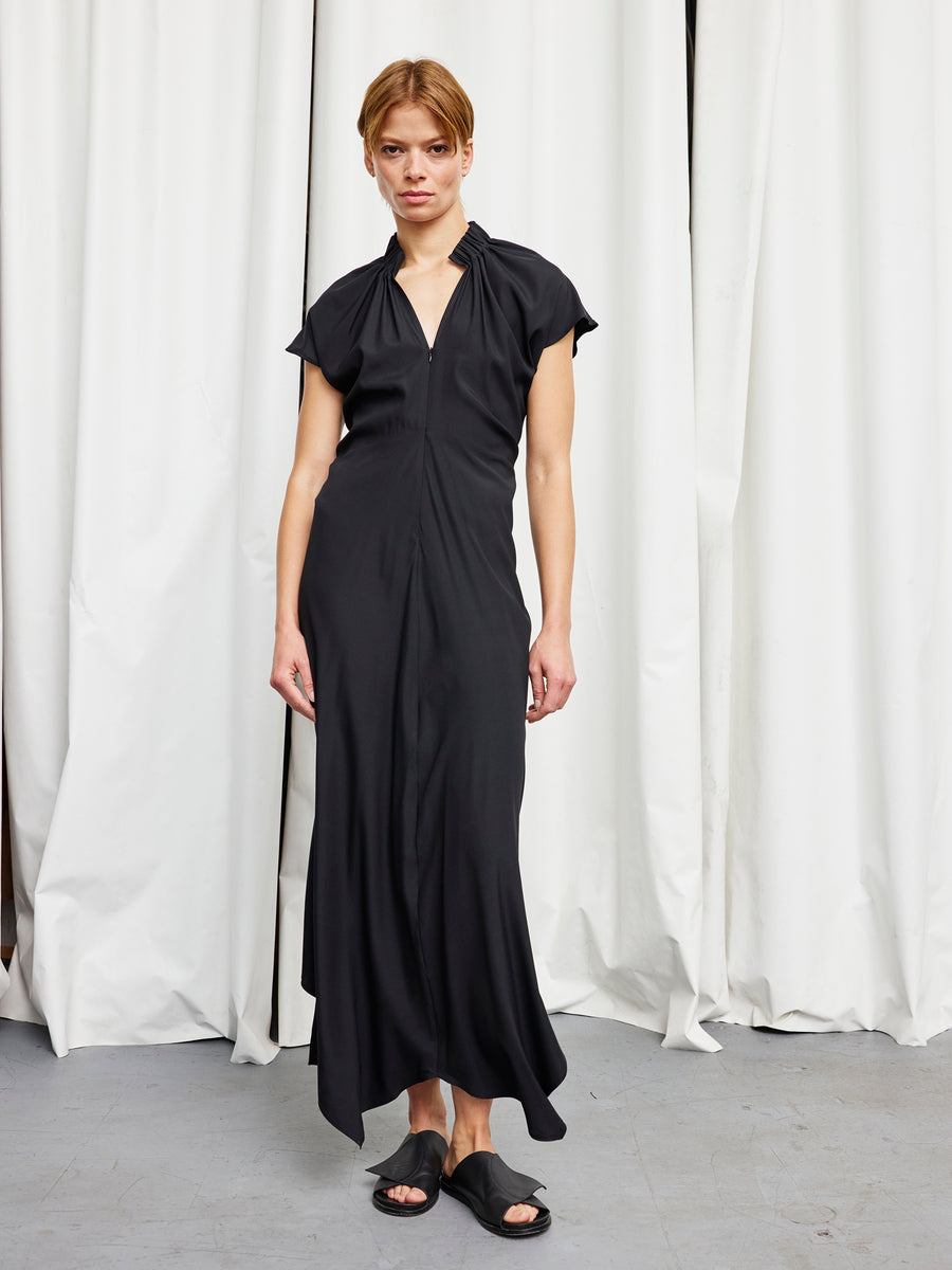A person stands indoors before white curtains, wearing the Long Ruched Stella Dress by Zero + Maria Cornejo and black slide sandals. The gray floor and flowing black silk dress create a minimalist, relaxed look.