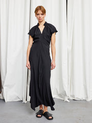 A person stands indoors before white curtains, wearing the Long Ruched Stella Dress by Zero + Maria Cornejo and black slide sandals. The gray floor and flowing black silk dress create a minimalist, relaxed look.