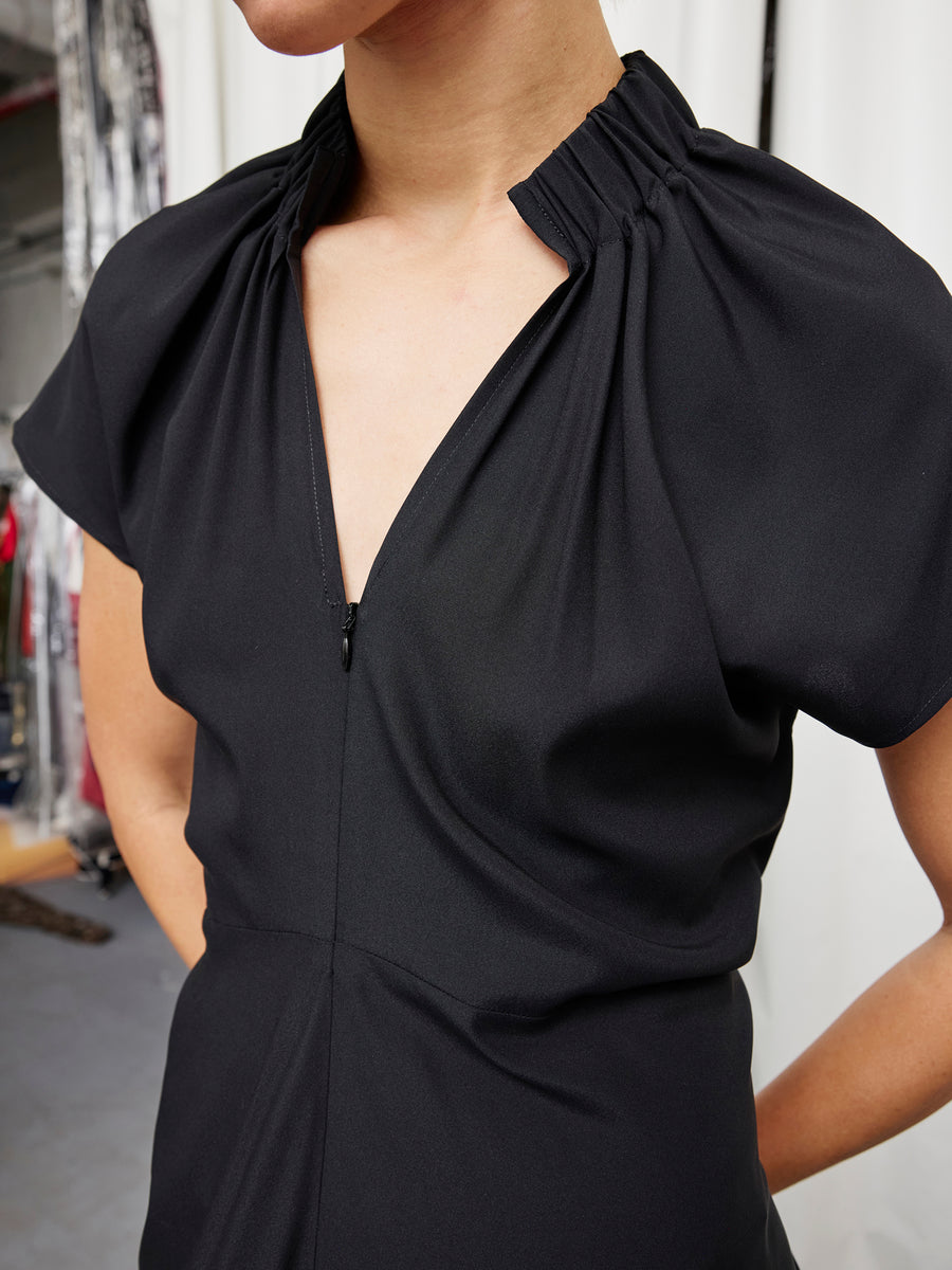 A person wearing the Long Ruched Stella Dress by Zero + Maria Cornejo—black stretch silk with gathered shoulders and a V-neckline—stands indoors against a blurred background.