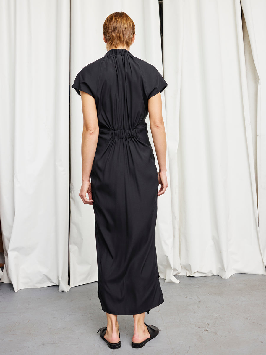 A person faces away, wearing the Zero + Maria Cornejo Long Ruched Stella Dress and black flats. They stand before white curtains on a light gray floor.