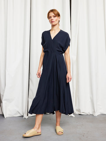A person wearing the Zero + Maria Cornejo Long Aki Wave Dress in navy blue with short sleeves and beige slip-on sandals stands before white curtains on a gray floor.