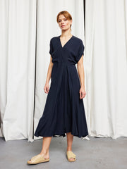 A person wearing the Zero + Maria Cornejo Long Aki Wave Dress in navy blue with short sleeves and beige slip-on sandals stands before white curtains on a gray floor.