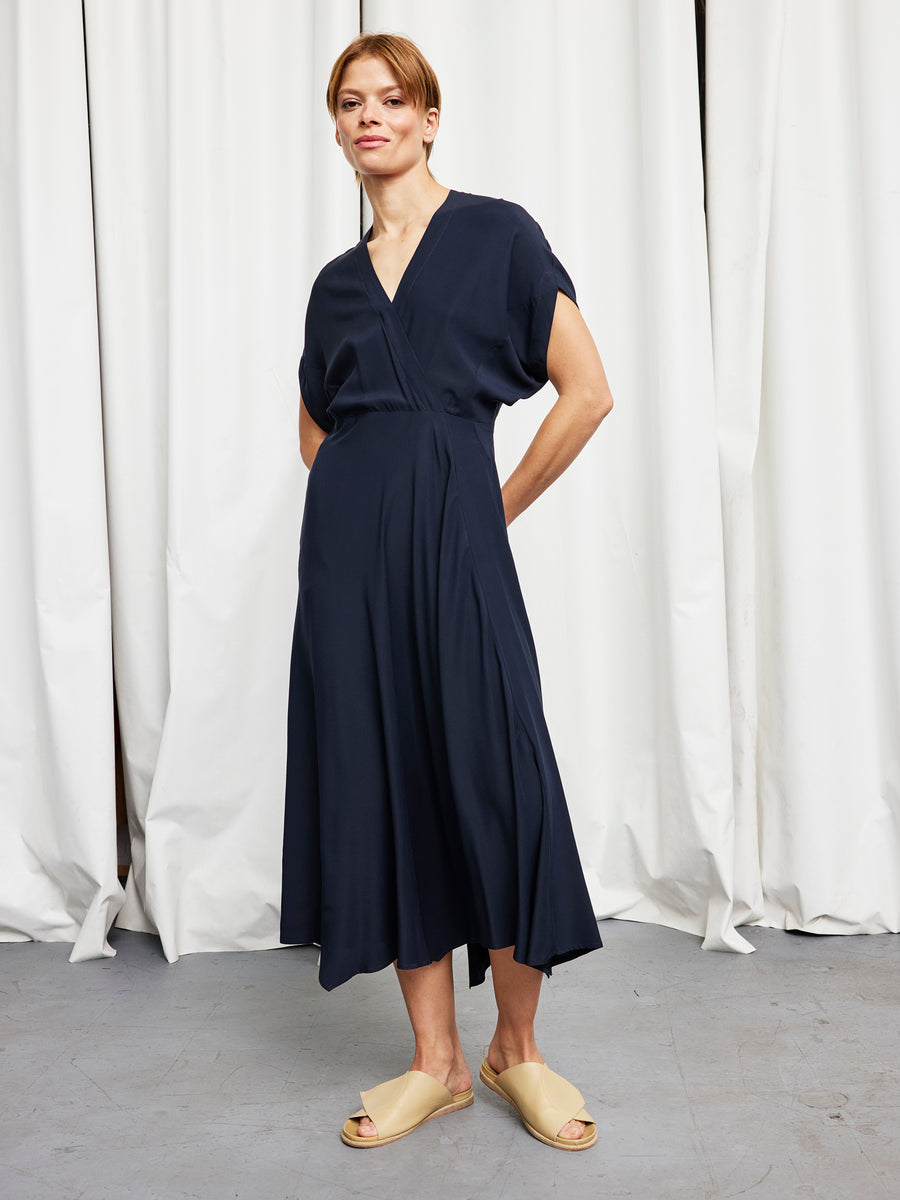 A person wears the Zero + Maria Cornejo Long Aki Wave Dress, posing indoors in front of white curtains on a grey floor.