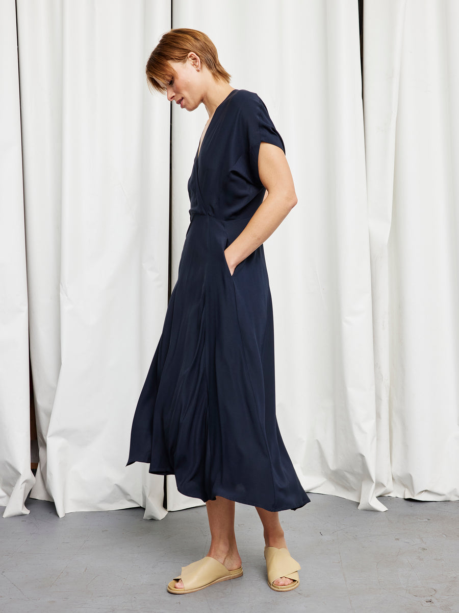 A person with short hair wears the Zero + Maria Cornejo Long Aki Wave Dress in loose navy silk charmeuse with short sleeves and pockets, standing sideways against white curtains.