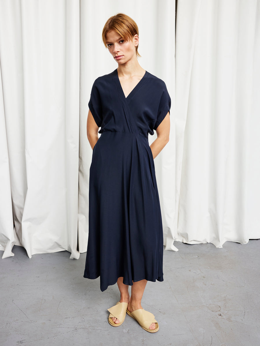 Someone stands on a gray floor before white curtains, hands in pockets and a neutral expression, wearing the Zero + Maria Cornejo Long Aki Wave Dress—a loose-fitting, short-sleeved, navy blue silk charmeuse dress—with beige sandals.