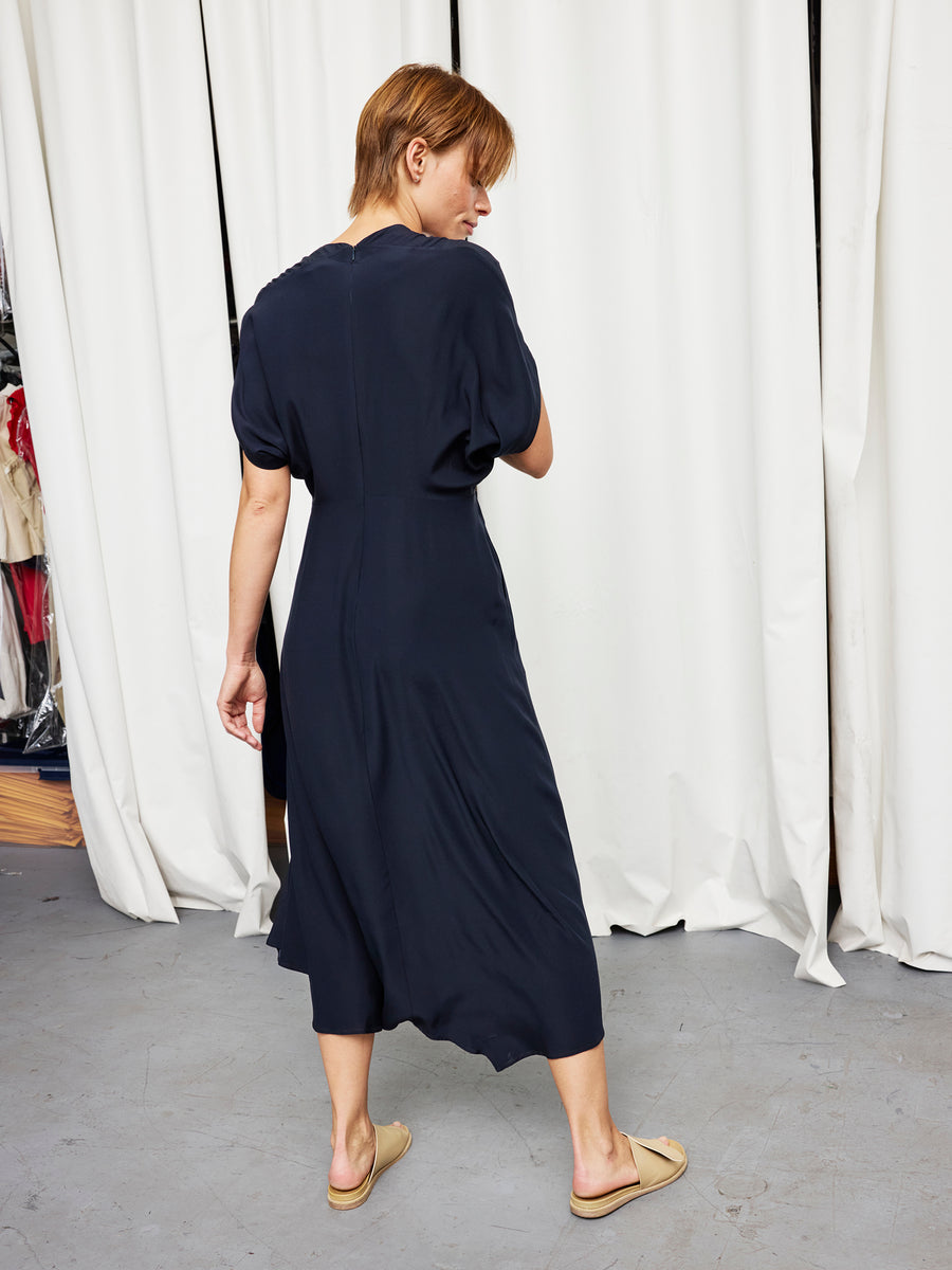 Someone with short hair stands indoors facing away from the camera, wearing the Zero + Maria Cornejo Long Aki Wave Dress— a loose-fitting, dark navy, full-length style—paired with beige slip-on shoes in front of white curtains.