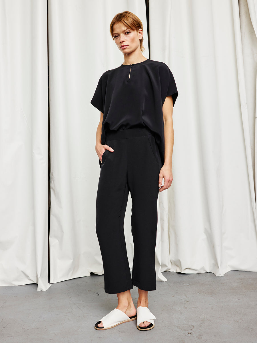 A person stands indoors against white curtains, wearing a loose black top and Zero + Maria Cornejo's Eko Pant, paired with white slide sandals. One hand is in their pocket and their expression is neutral.