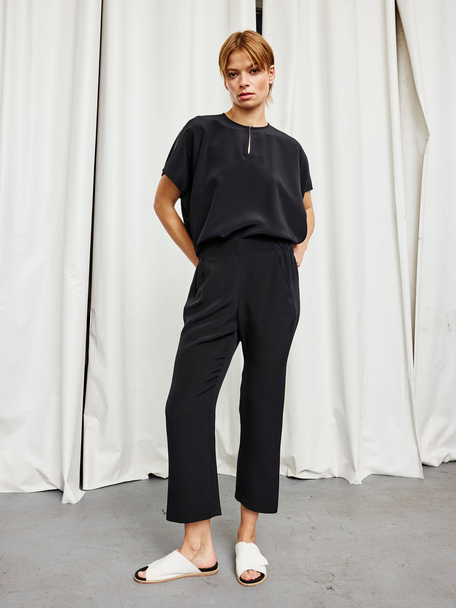 A person stands indoors before white curtains, wearing the black woven silk Eko Pant by Zero + Maria Cornejo, a loose black top, and white slide sandals. They have short light brown hair and a neutral expression with hands in their pockets.