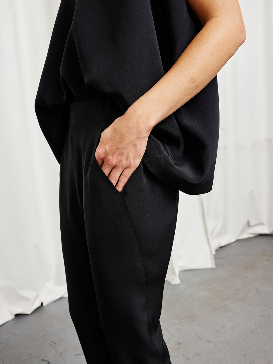 A person models the Eko Pant by Zero + Maria Cornejo—loose-fitting woven silk pants—paired with a black top, standing with one hand in their pocket before white curtains and a gray floor.
