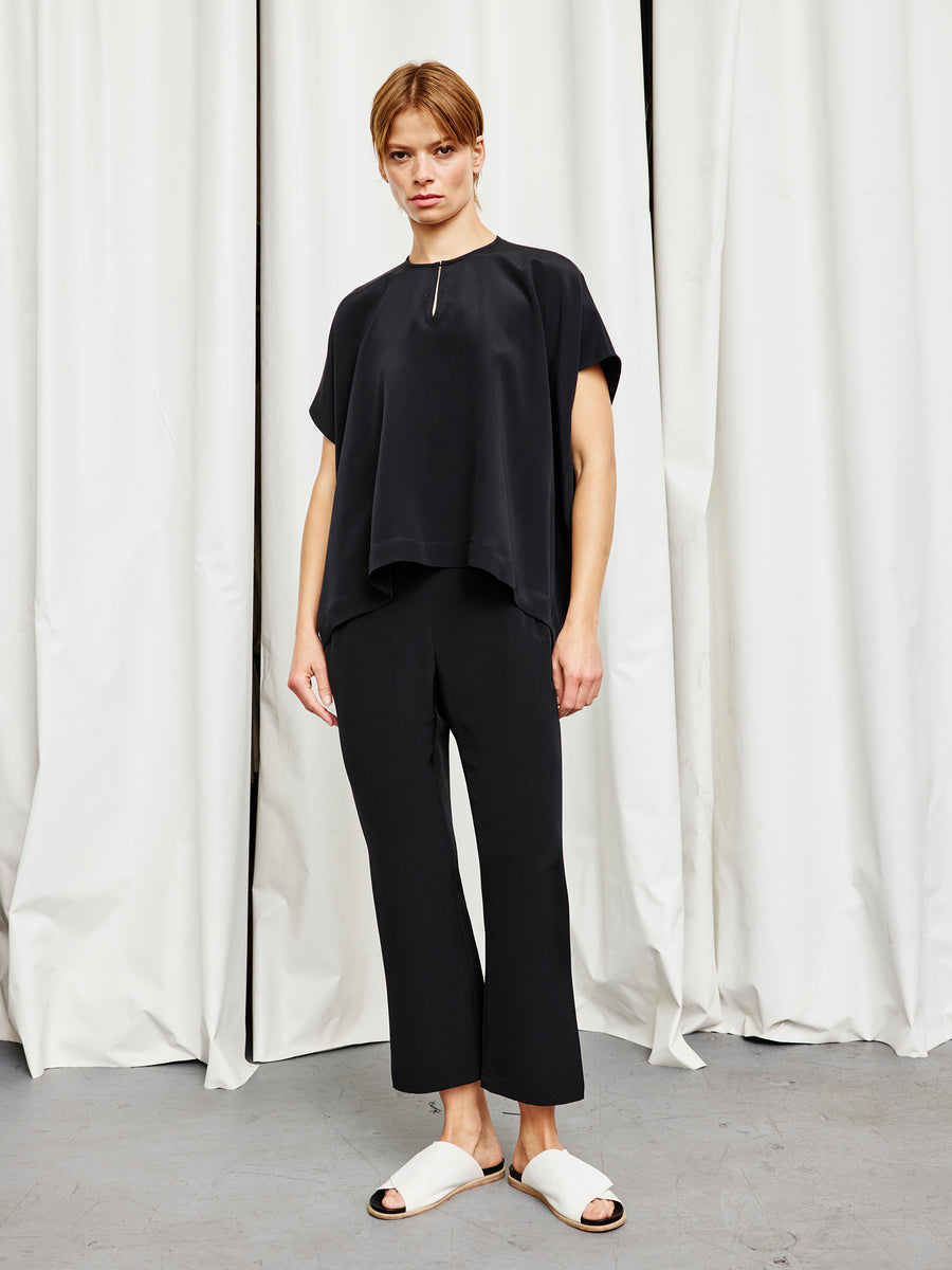 A person stands indoors against white curtains, wearing the Rae Poncho Top by Zero + Maria Cornejo, black pants, and white slide sandals. They look at the camera with a neutral expression, embodying effortless sustainable fashion.