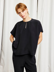A person with short light brown hair wears the Rae Poncho Top in black by Zero + Maria Cornejo, paired with matching pants and standing with hands in pockets before a light curtain backdrop—a nod to sustainable fashion.