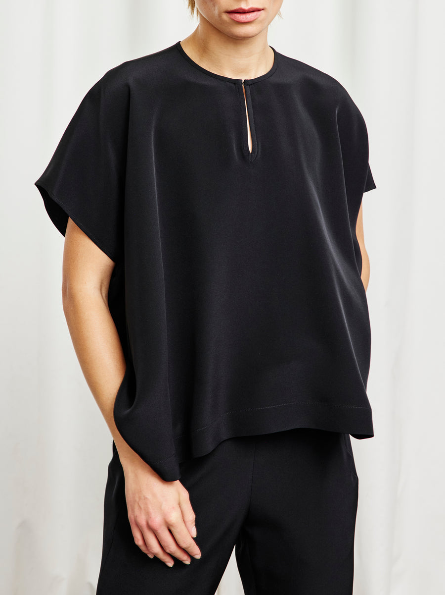 Person wears the Rae Poncho Top by Zero + Maria Cornejo—a loose black silk piece with wide sleeves and a front keyhole—paired with black pants, standing against a white background in timeless sustainable fashion.