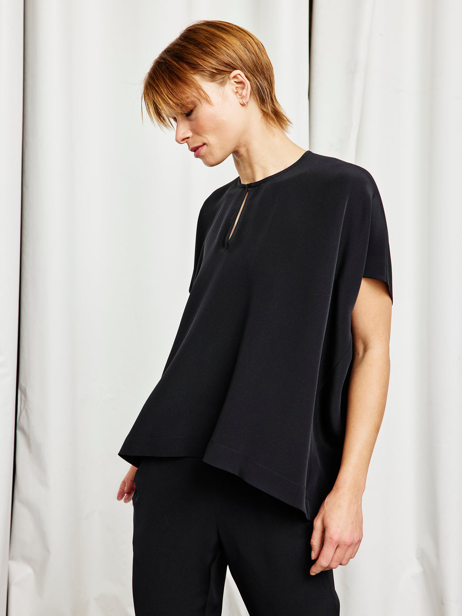 Someone with short, light brown hair wears the Zero + Maria Cornejo Rae Poncho Top and matching black pants, standing against a white curtain and looking downward with a neutral expression.