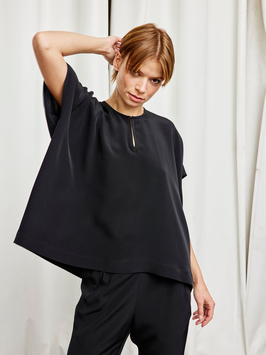 A person with short light brown hair poses with one hand behind their head in front of white curtains, wearing the Rae Poncho Top by Zero + Maria Cornejo and black pants.