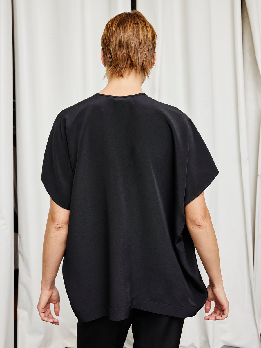A person with short, light brown hair stands with their back to the camera in front of white curtains, wearing the Zero + Maria Cornejo Rae Poncho Top and black pants, showcasing sustainable fashion.