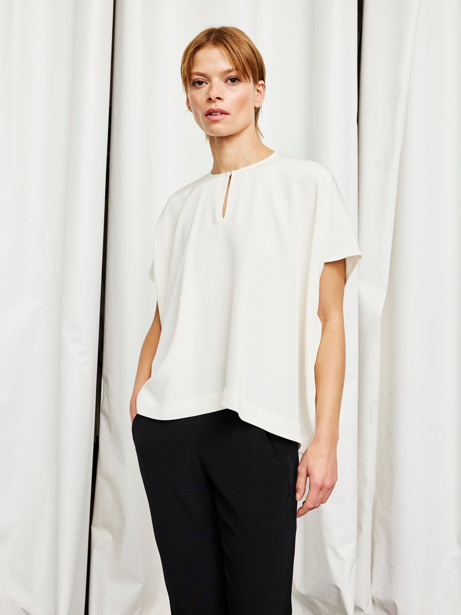 A person stands against white curtains, wearing the Zero + Maria Cornejo Rae Poncho Top—a loose-fit, woven silk piece with a front keyhole and short sleeves—paired with black pants, embodying effortless sustainable style.