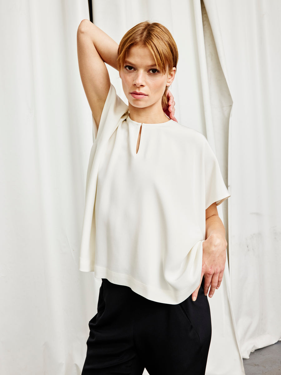 A woman stands before white curtains in the Rae Poncho Top by Zero + Maria Cornejo, color White Pepper, and black pants. With one hand behind her head and a serious look, she embodies effortless, sustainable style.