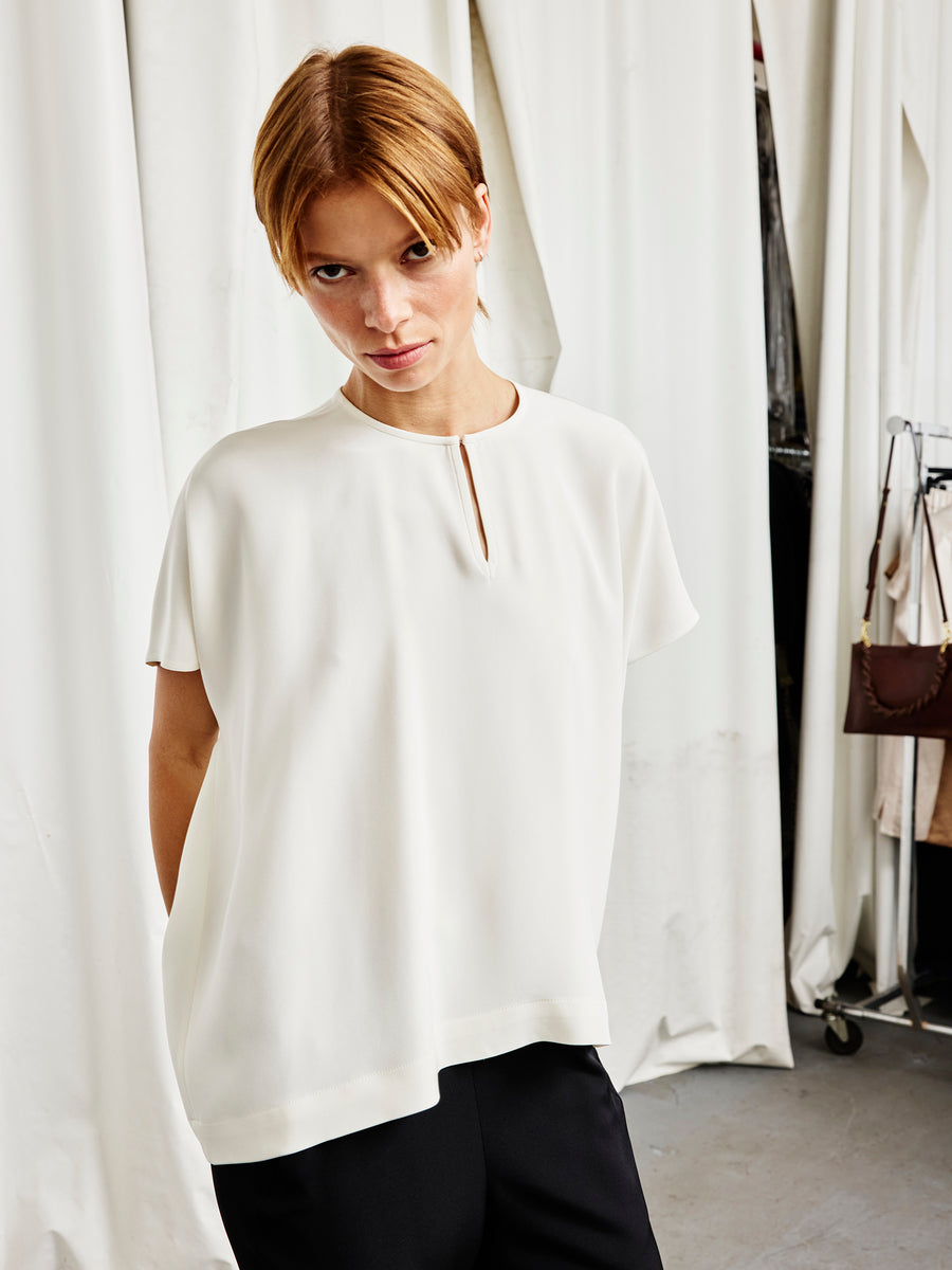 A person with short, light brown hair stands indoors wearing the Zero + Maria Cornejo Rae Poncho Top—a loose, white woven silk style with a small keyhole neckline—paired with black pants. White curtains and clothing racks are in the background.