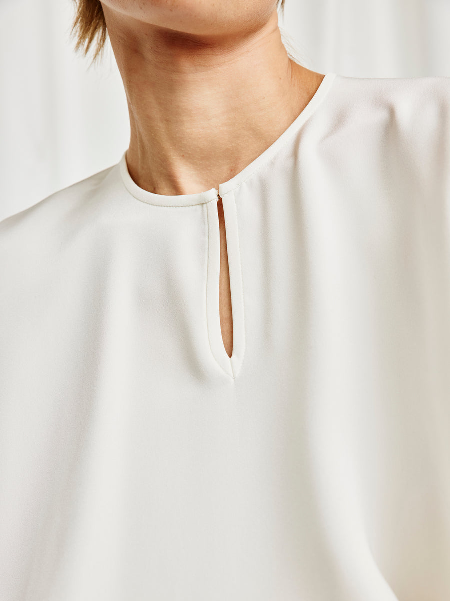 Close-up of a person wearing the Rae Poncho Top by Zero + Maria Cornejo, featuring a small vertical keyhole neckline—an elegant, sustainable fashion choice—against a softly blurred light background.
