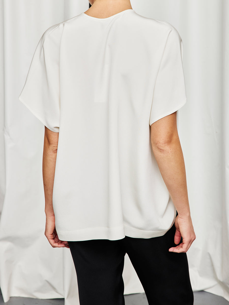 A person stands with their back to the camera, wearing the Rae Poncho Top by Zero + Maria Cornejo and black pants, against a white curtain background, highlighting effortless style and sustainable fashion.