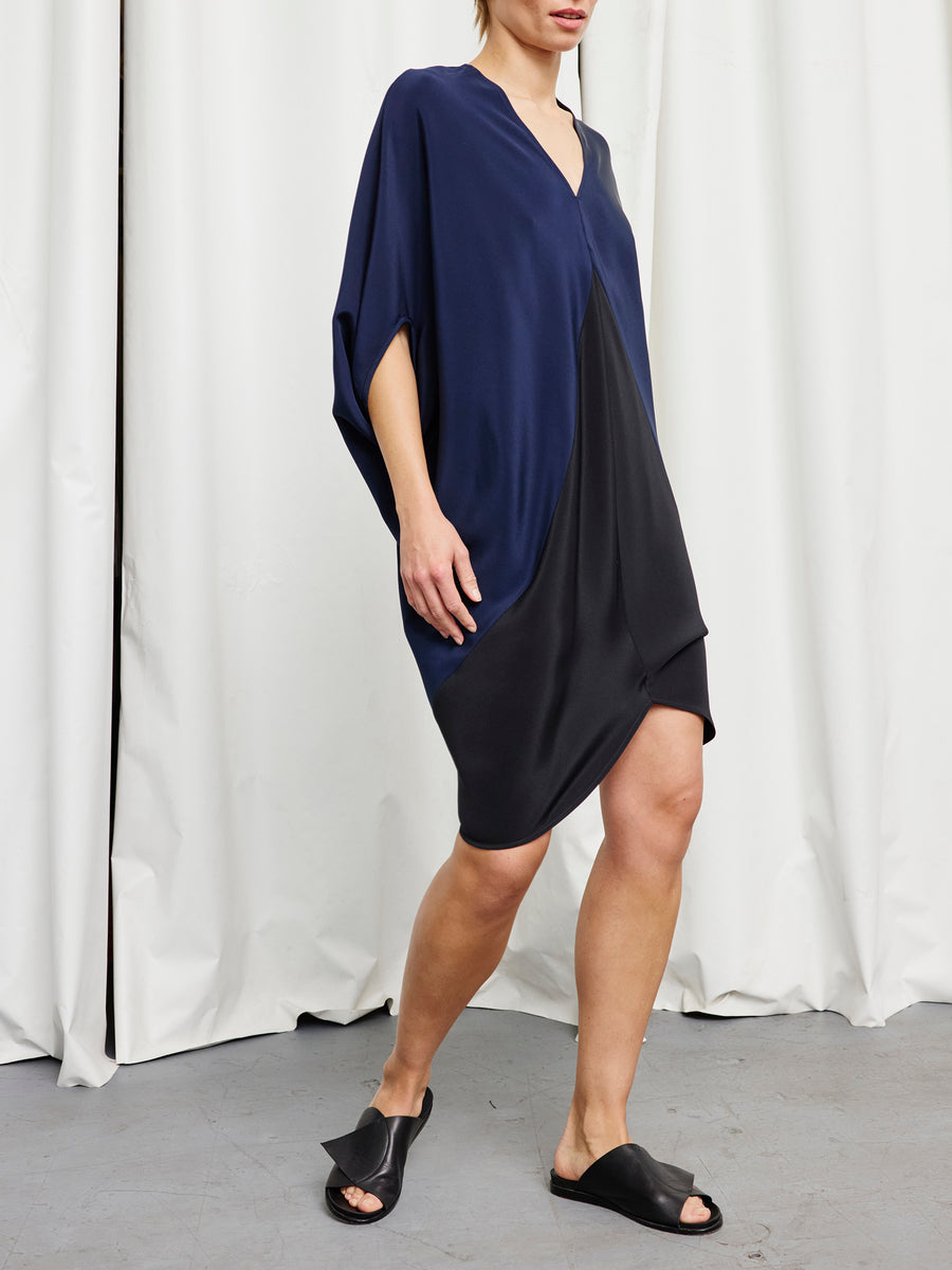 A person stands on a gray floor before white curtains, wearing the Zero + Maria Cornejo Koya Dress—a loose, navy and black color-blocked cocoon silhouette with short sleeves—paired with black slide sandals. Their face is partially out of frame.