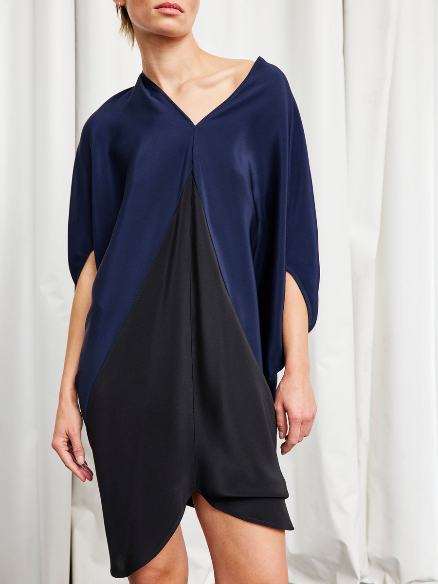 A person stands against a white curtain wearing the Koya Dress by Zero + Maria Cornejo, a loose-fitting silk crepe v-neck dress with navy top and black panels, three-quarter sleeves, and a cocoon silhouette. Their face is partially out of frame.