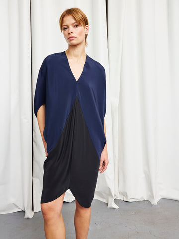 A woman with short light brown hair and a neutral expression stands before white curtains, wearing the Koya Dress by Zero + Maria Cornejo—a chic v-neck style with a navy top draped over a fitted black skirt, one hand in her pocket.