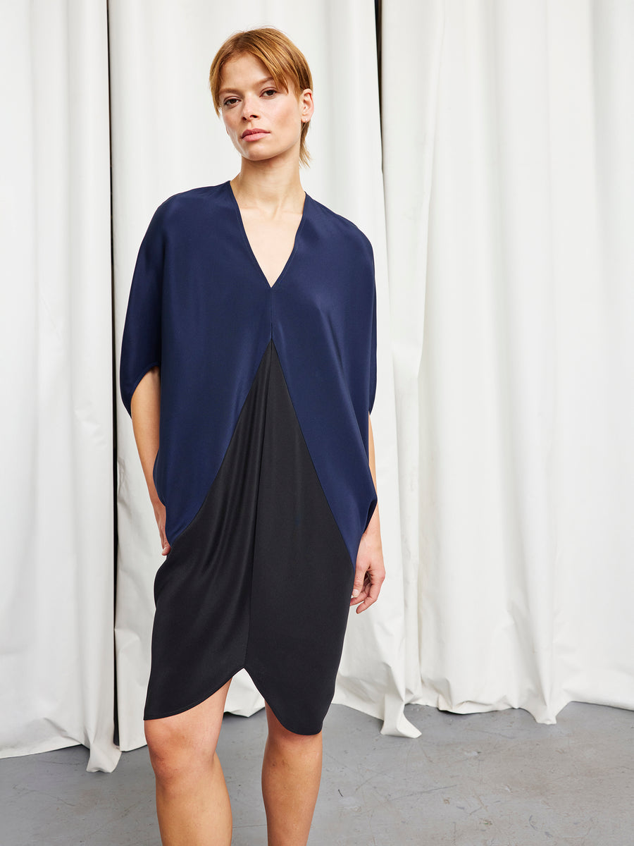 A woman with short light brown hair and a neutral expression stands before white curtains, wearing the Koya Dress by Zero + Maria Cornejo—a chic v-neck style with a navy top draped over a fitted black skirt, one hand in her pocket.