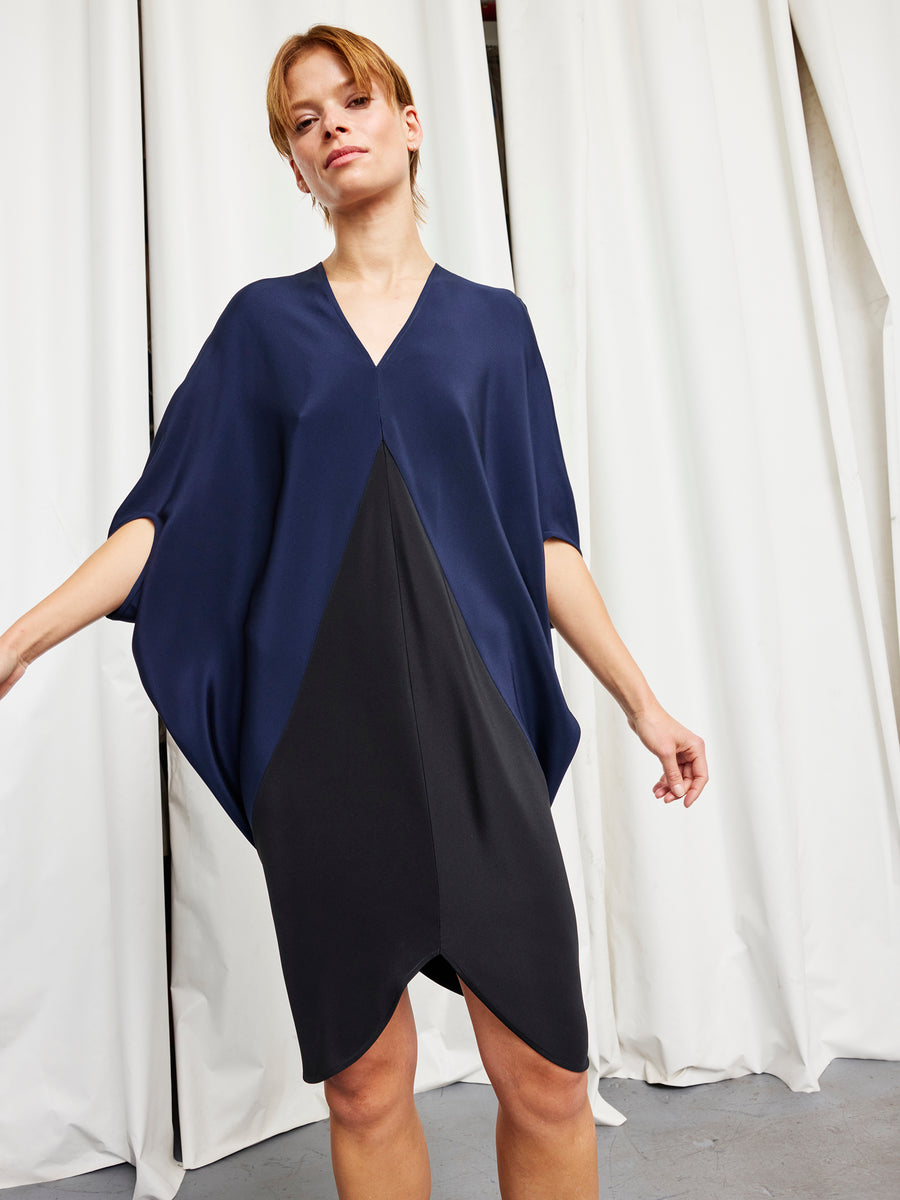 A person wears the Koya Dress by Zero + Maria Cornejo—a loose-fitting, navy and black color-blocked v-neck with batwing sleeves and a cocoon silhouette—standing before white curtains on a gray floor.