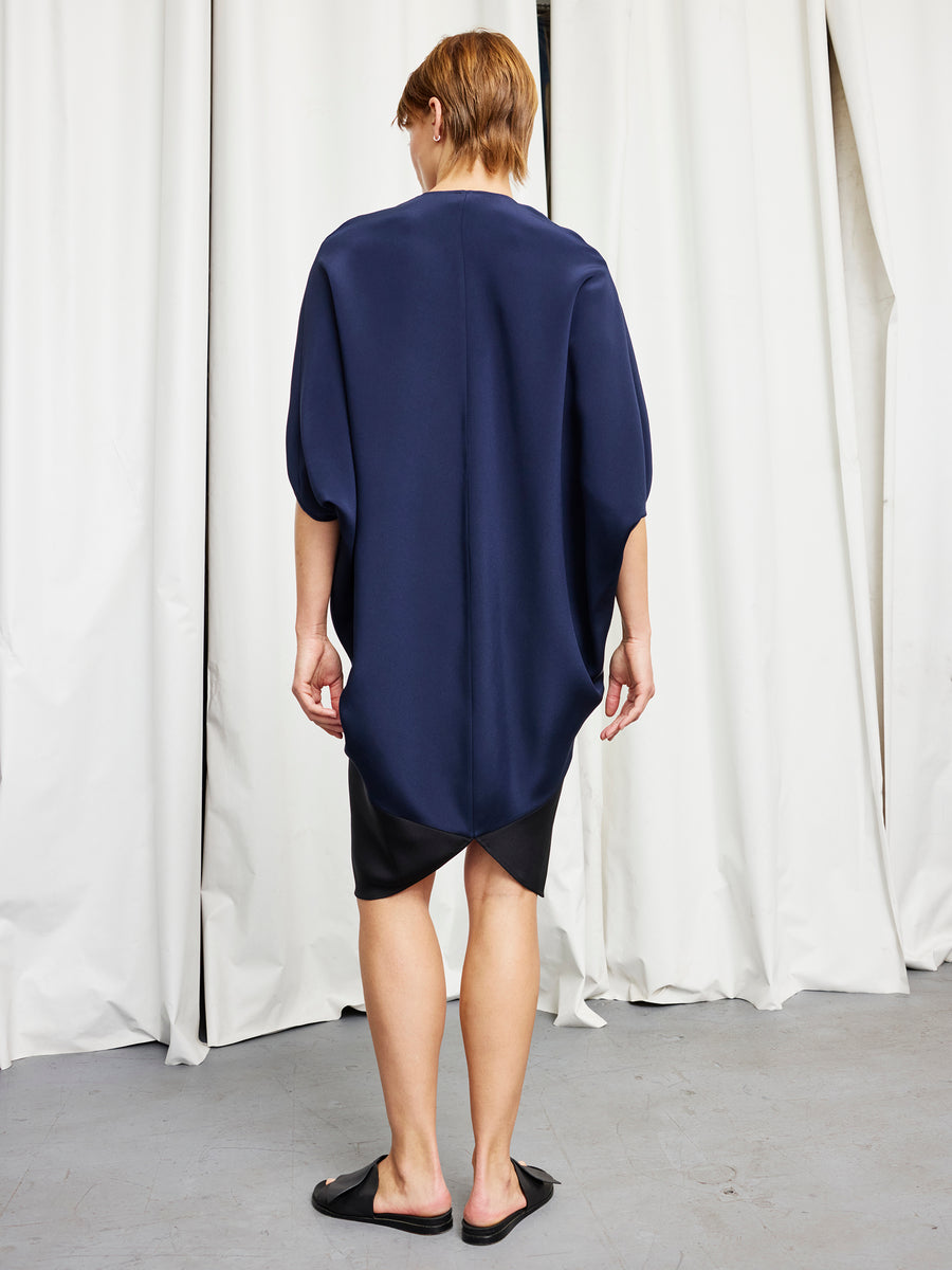 A person stands indoors with their back to the camera, wearing the navy Koya Dress by Zero + Maria Cornejo, featuring a cocoon silhouette. White curtains are visible in the background.