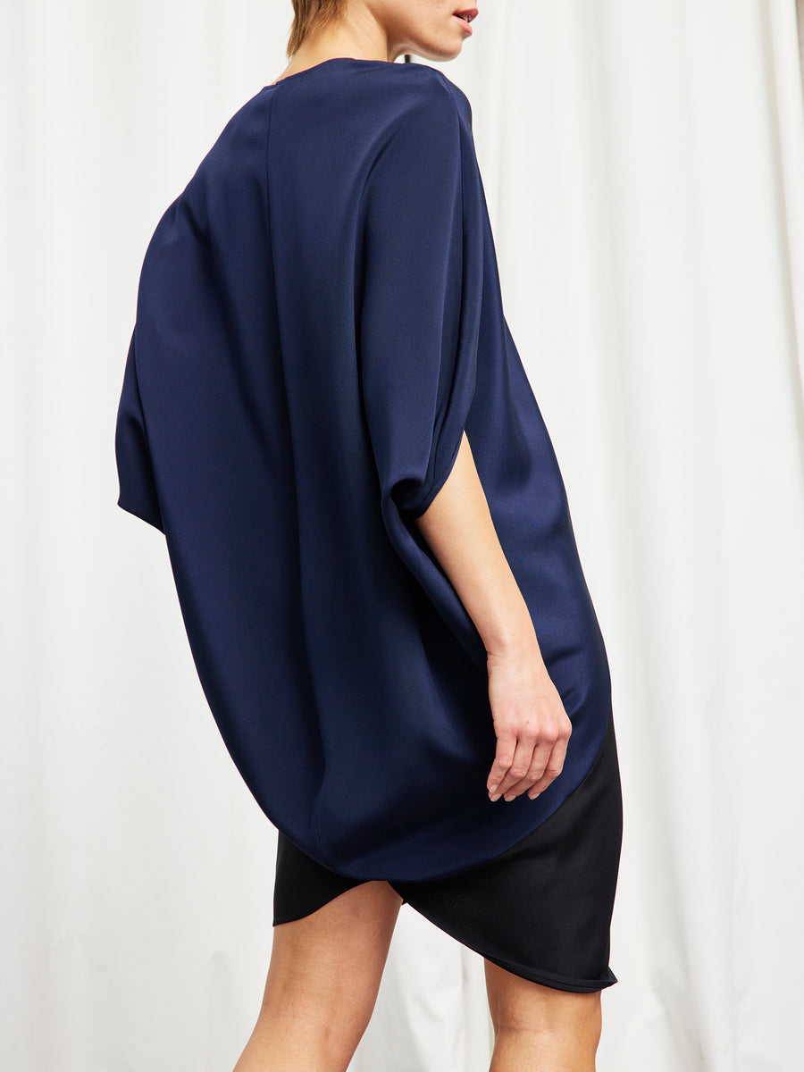 A person stands in profile before a white curtain wearing the Zero + Maria Cornejo Koya Dress, a loose-fitting navy silk crepe piece with wide sleeves, an asymmetrical hem, and a soft cocoon silhouette.