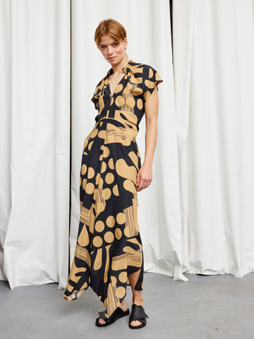 A person stands before white curtains wearing the Long Ruched Stella Dress by Zero + Maria Cornejo, a black and tan patterned silk charmeuse gown with a deep V-neck, front slit, and black open-toed shoes for an effortlessly elegant look.