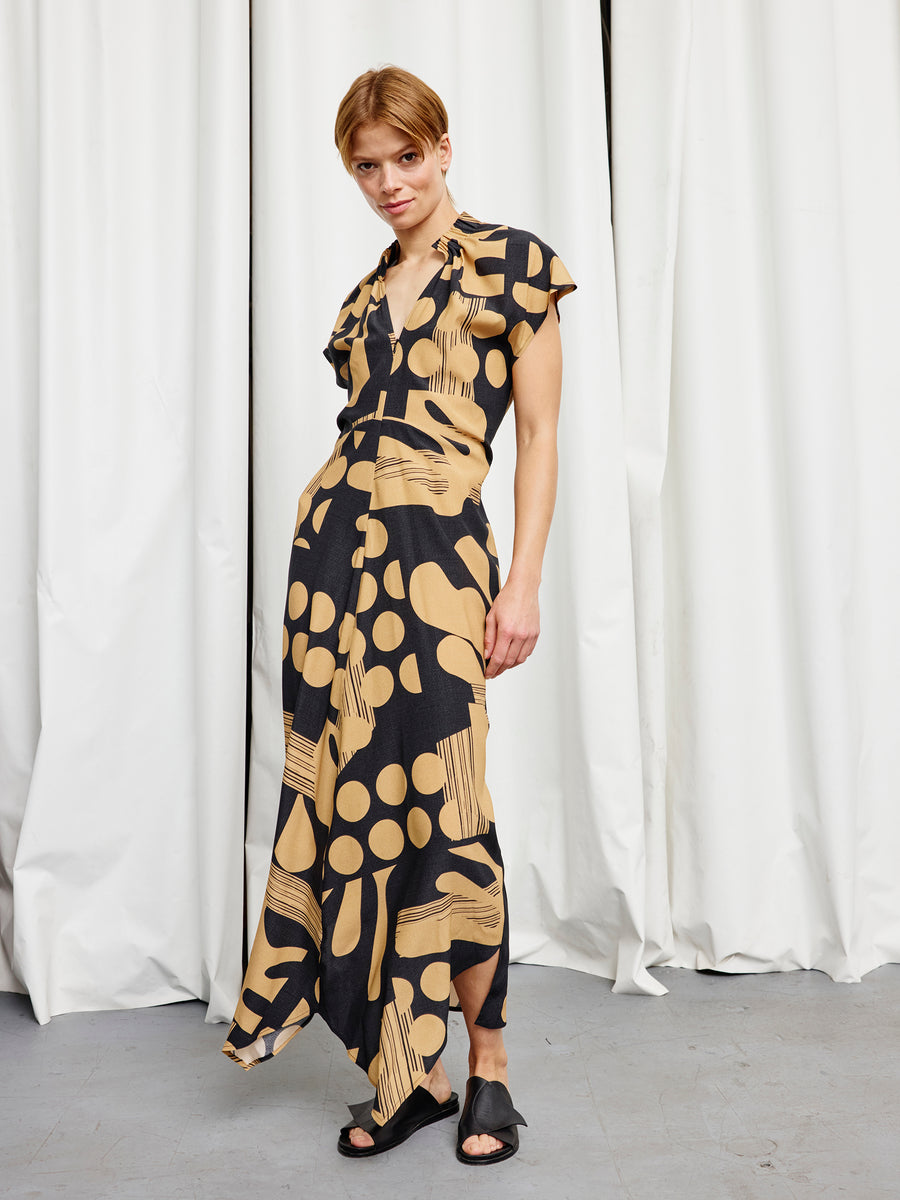 A person stands before white curtains wearing the Long Ruched Stella Dress by Zero + Maria Cornejo, a black and tan patterned silk charmeuse gown with a deep V-neck, front slit, and black open-toed shoes for an effortlessly elegant look.