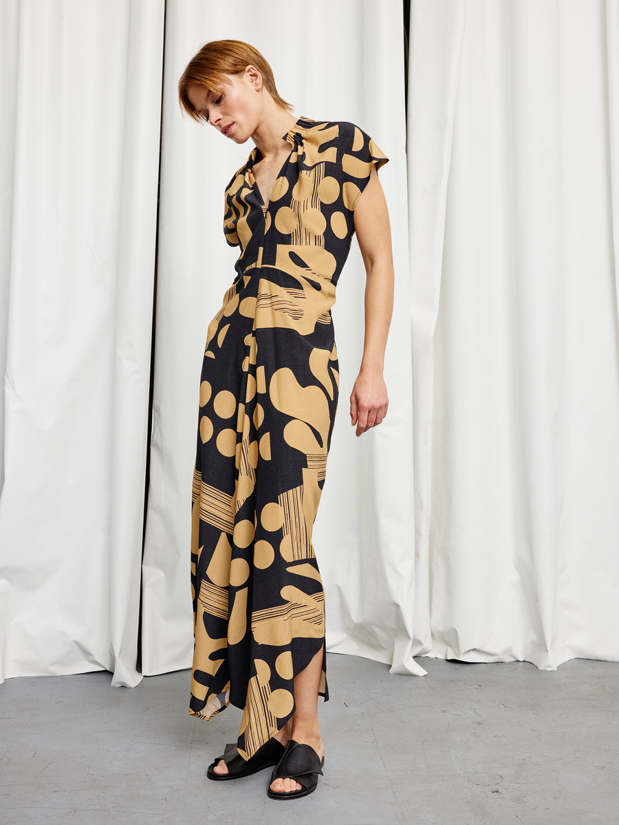 A person stands indoors in the Long Ruched Stella Dress by Zero + Maria Cornejo, featuring tan abstract shapes and dots on black silk charmeuse, paired with black slide sandals and set against white curtains.