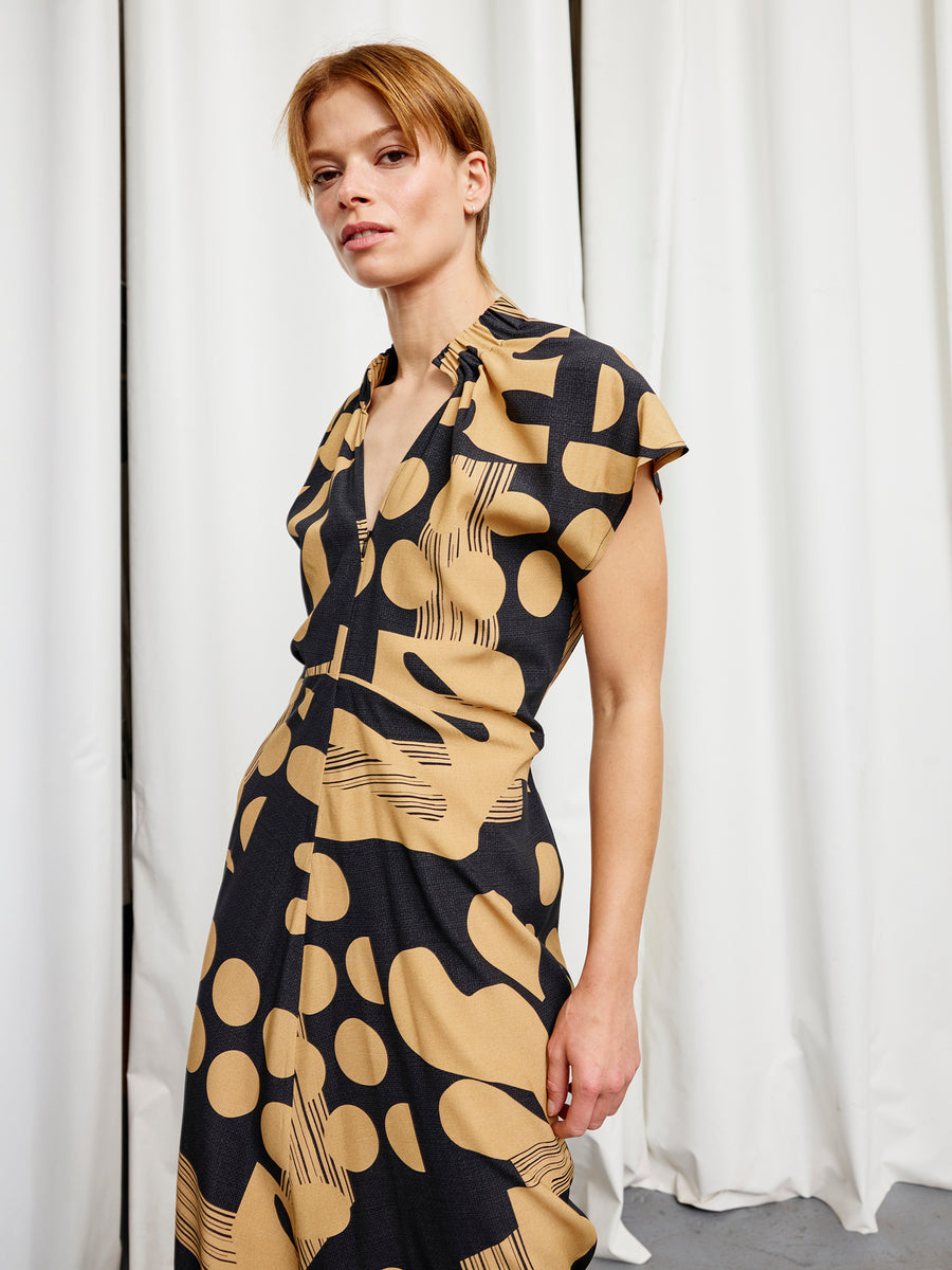 A person with short light brown hair wears the Long Ruched Stella Dress by Zero + Maria Cornejo—featuring short sleeves and a beige and black pattern—while standing in front of white curtains.