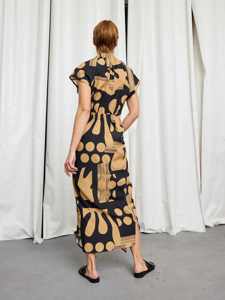A person stands facing away, wearing the Long Ruched Stella Dress by Zero + Maria Cornejo—a black and tan patterned silk charmeuse, short-sleeve dress with a gathered waist. The full-length design flows to the ankles above black sandals on a gray floor before white curtains.