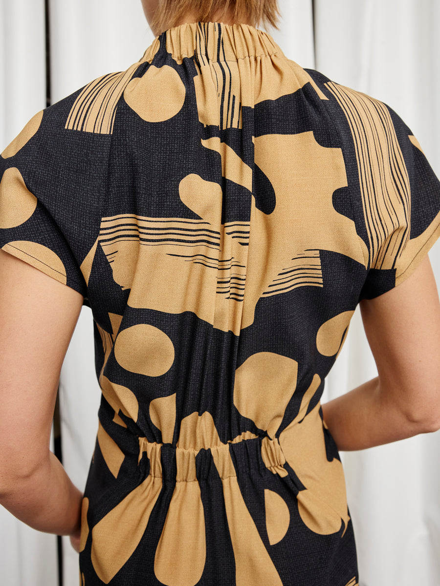 A person is shown from the back wearing the Long Ruched Stella Dress by Zero + Maria Cornejo, a short-sleeved, full-length dress with a gathered waist and high neckline in an abstract black and tan dot-and-line print. Made in New York.