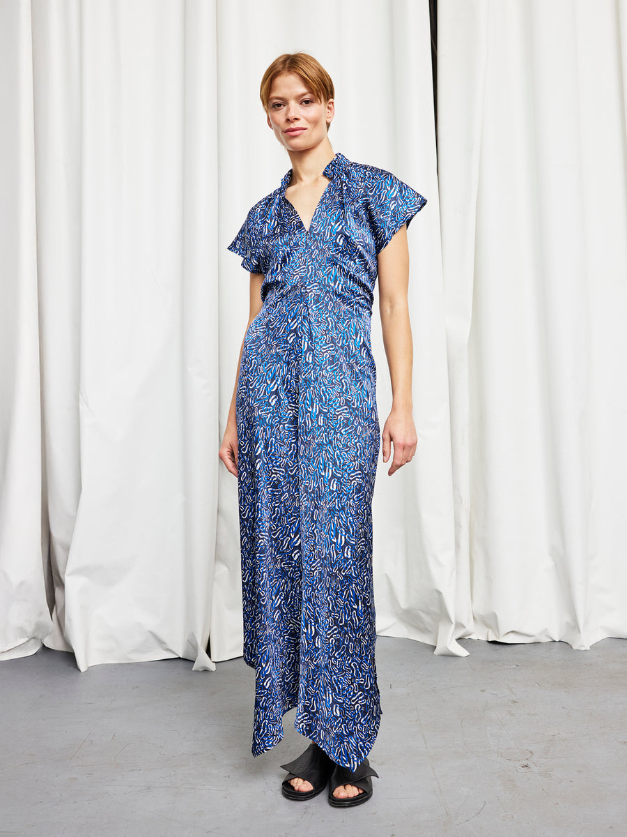 A person with short light brown hair and a neutral expression stands indoors before white curtains, wearing the Zero + Maria Cornejo Long Ruched Stella Dress in blue silk charmeuse and black sandals.