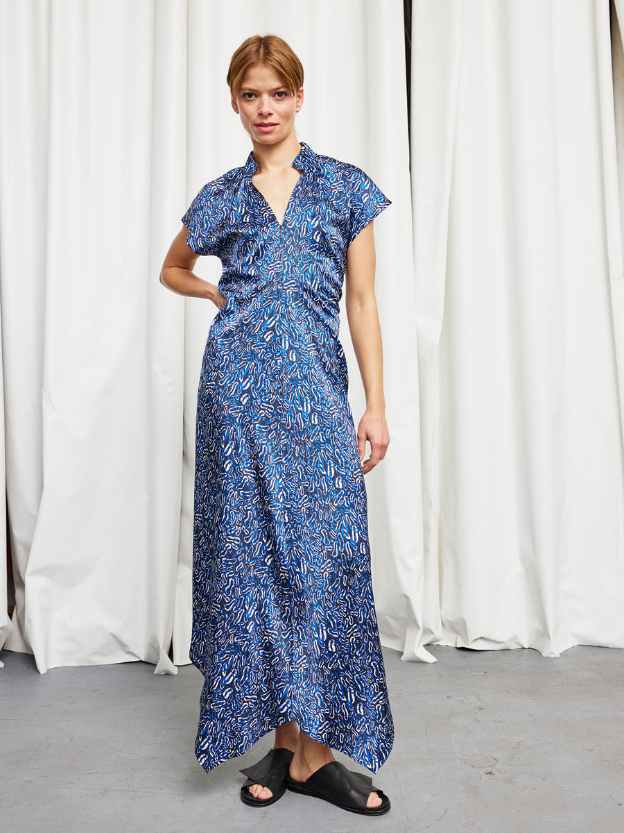 A person with short hair poses indoors by white curtains, wearing the Zero + Maria Cornejo Long Ruched Stella Dress in blue silk charmeuse and black slip-on sandals, with one hand on their hip.